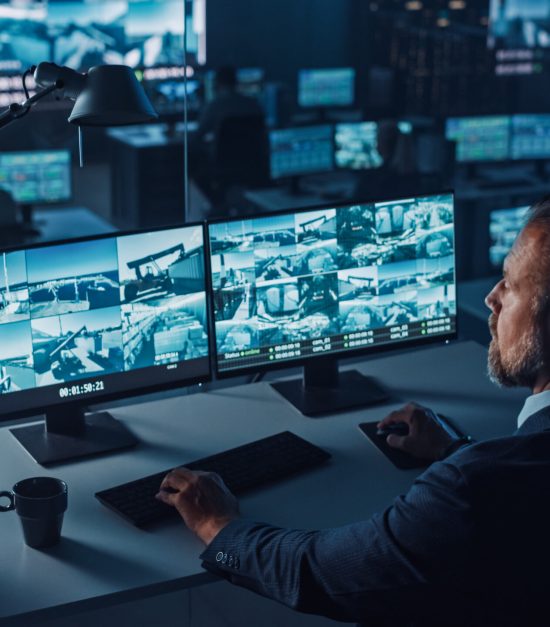 Male Officer Works on a Computer with Surveillance CCTV Video Footage in a Harbour Monitoring Center with Multiple Cameras on a Big Digital Screen. Employees Sit in Front of Displays with Big Data.